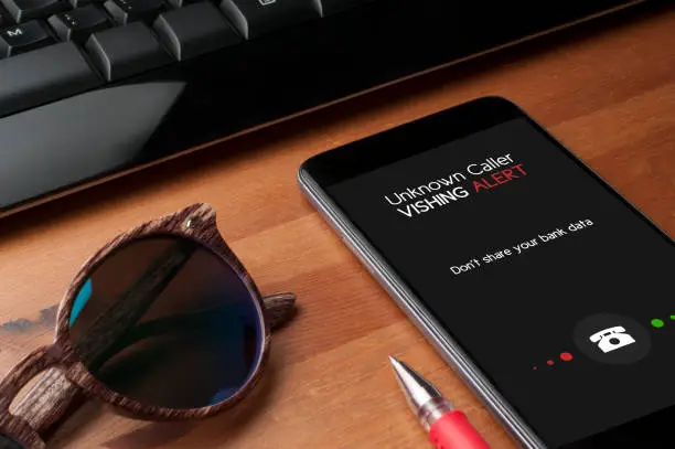 Vishing (voice phishing) concept, a smartphone on a table next to a computer keyboard and a pair of sunglasses show an unknower caller call with vishing alert and a reminder to not share personal bank data