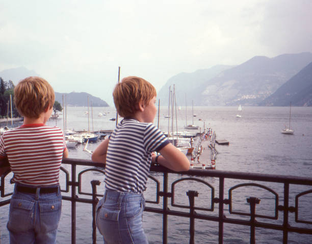 Vintage 1982 omage of two young boys both wearing jeans shorts and striped t-shirts overlooking lake Geneva in Switzerland. Vintage 1982 analog camera image of two young boys / brothers seen from behind both wearing jeans shorts and striped t-shirts overlooking lake Geneva or Lac Léman from a balcony in Switzerland. sailing photos stock pictures, royalty-free photos & images
