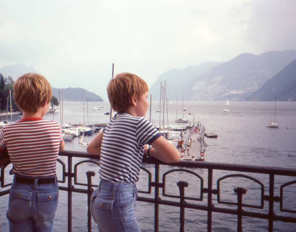 vintage 1982 fabricante de dois meninos novos que desgastam shorts das calças de brim e t-shirt listrados que negligenciam o lago genebra em switzerland. - 1980s style fotos - fotografias e filmes do acervo