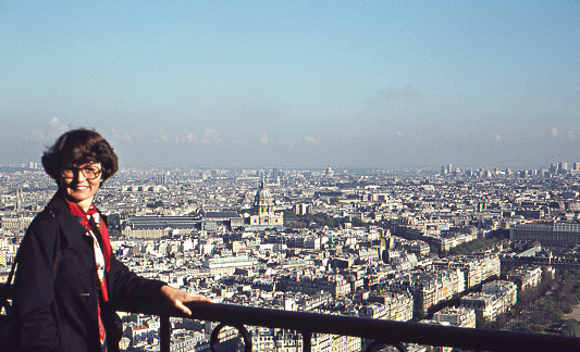 Vintage 1975 analog image of a young woman in a blue coat and red scarf enjoying the view and posing on the Eiffel tower with Paris in the background.