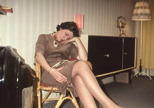 Dutch 1958 vintage image of a woman with magazine on lap sleeping in chair next to an old gas heater.
