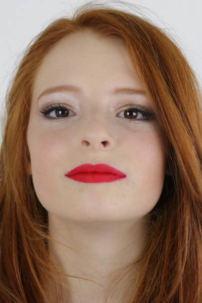 Stock photo showing a close-up of a beautiful, ginger haired teenager modelling a glam makeup look wearing bright red lipstick. imagen de adolescente pelirroja adolescente 14 / 15 con piel pálida y pecas se sentó en el dormitorio probando el tono rojo brillante de lápiz labial en sus labios, puchero labial rodeado de luces de cuerda de hadas, retrato de maquillaje facial natural