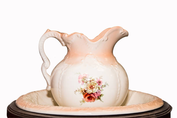 An old-fashioned ceramic water pitcher and wash basin resting on a marble countertop. The antique pottery vessel, decorated with cream color glaze and curving leaf stem pattern,  reflects a pioneer era without indoor plumbing and running water for washing up. Horizontal, quaint still life in warm tones, with copy space.
