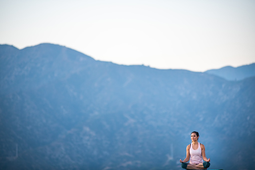 A young hispanic woman practicing yoga appears to be floating in the bottom right corner of the frame. She sits cross legged in the Sukhasana position with a larger than life mountain range behind her.