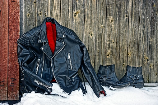 black leather jacket and boots stand on white snow against a gray wooden wall