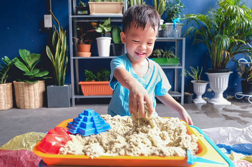 Cute smiling Asian 3 - 4 years old toddler boy playing with kinetic sand in sandbox at home / nursery / day care, Fine motor skills development, Montessori education, Creative play for kids concept