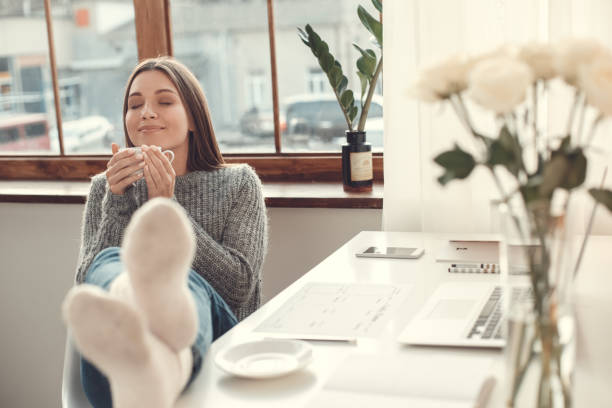 Young woman freelancer indoors home office concept winter atmosphere drinking coffee relaxation Young female freelancer at home office winter sitting smiling smell the coffee relaxation side view feet up stock pictures, royalty-free photos & images