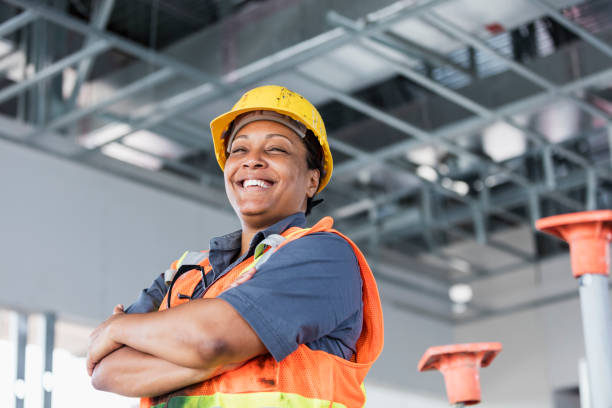 Female African-American construction worker A mature African-American woman in her 40s wearing a hardhat and reflective vest, a construction worker or building contractor, standing with her arms crossed, smiling at the camera. role reversal stock pictures, royalty-free photos & images