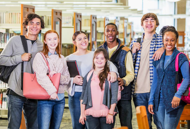 High school students in library, girl with down syndrome A multi-ethnic group of seven high school students, 15 to 17 years old, standing together in a library, smiling and looking at the camera. The girl in the middle has down syndrome. down syndrome photos stock pictures, royalty-free photos & images