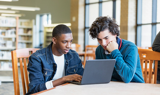 Two multi-ethnic teenage boys studying together, sitting at a table in the library. They are 16 year old high school students. The African-American student is typing on his laptop while his Hispanic friend watches.