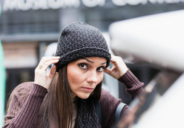 Close-up of a mature mixed race Hispanic and Caucasian woman in her 40s with a serious expression, outdoors in the city. She is dressed warmly, for autumn, in a sweater, adjusting the knit hat on her head, as she looks toward the camera.