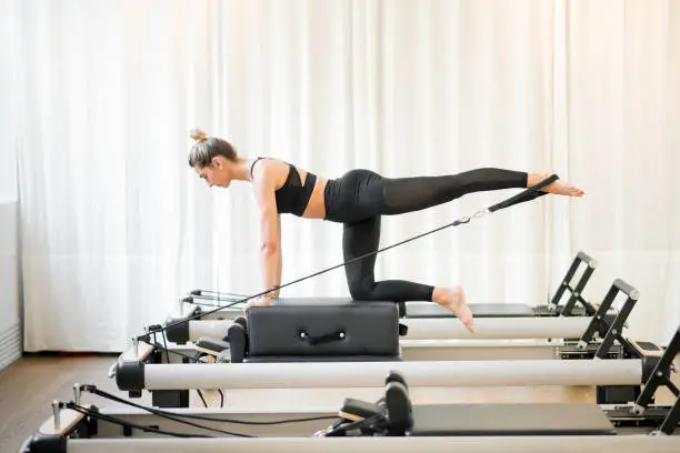 Woman performing a pilates diagonal stabilisation exercise using a strap on a reformer bed in a gym in a health and fitness concept