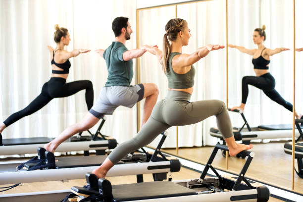 Class in a gym doing pilates standing lunges Class in a gym doing pilates standing lunges on reformer beds to stretch and tone the muscles reflected in a wall mirror pilates photos stock pictures, royalty-free photos & images
