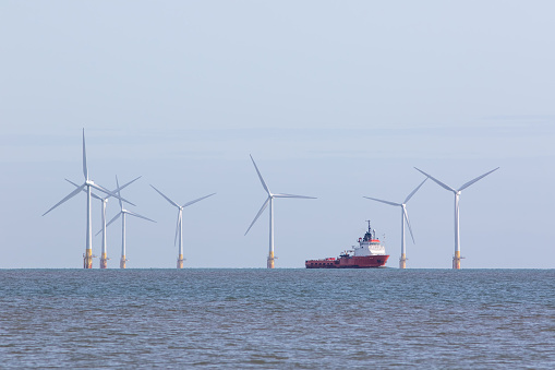 Renewable energy offshore wind farm turbines with passing maintenance supply vessel ship. Windfarm on the sea horizon with boat sailing between towers. Sustainable resource green power development.