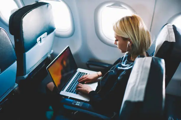 Photo of Attractive caucasian female passenger of airplane sitting in comfortable seat listening music in earphones while working at modern laptop computer with mock up area using wireless connection on board