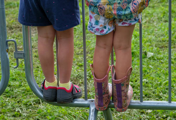 View from behind of two children wearing bright clothing, standing on a metal barrier at a sporting event. They have climbed onto the bottom bar to be able to get a better view. The young boy is wearing blue shorts, yellow socks and grey and red sports shoes. The young girl is wearing colourful shorts with a fruit pattern and a colourful jacket and pink and purple rubber boots.