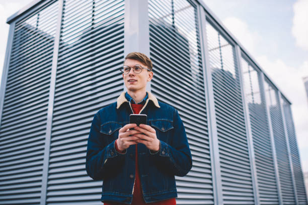 Pondering young male blogger in stylish denim jacket looking away while messaging in chat on smartphone using 4G internet standing in downtown.Contemplative hipster guy installing app on cellular reflexionando joven blogger masculino con elegante chaqueta de mezclilla mirando hacia otro lado mientras que la mensajería en el chat en el teléfono inteligente usando internet 4g de pie en el centro de la ciudad. contemplado hipster chico instalando ap