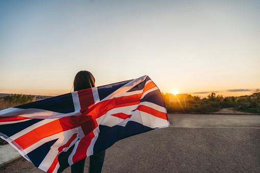 Young woman spreading British flag outdoors, while sun is going down. Rear view.