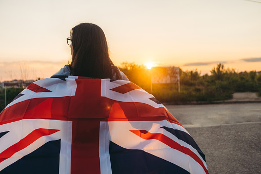 Young woman spreading British flag outdoors, while sun is going down. Rear view.