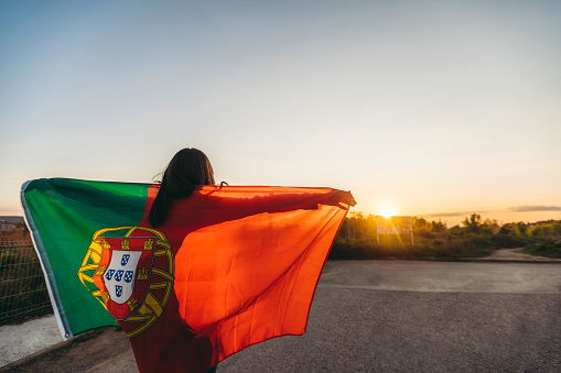 Young woman spreading Portuguese flag outdoors, while sun is going down. Rear view.