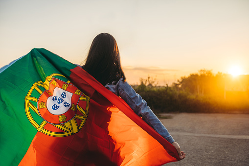 Young woman spreading Portuguese flag outdoors, while sun is going down. Rear view.