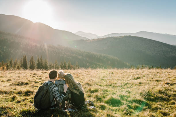 felice famiglia hipster: mamma, papà, figlia con zaino che si gode il tramonto sulla cima della montagna nebbiosa. turisti viaggiatore in background vista mockup. escursionisti alla ricerca della luce del sole in viaggio nel paese europa. - carpathian mountain range adventure mountain peak mountain foto e immagini stock