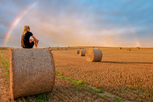 Young woman sitting on a hay bale while overlooking the landscape during harvest season.