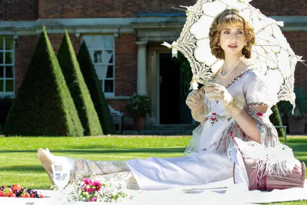 Photo of Beautiful lady in regency clothing sitting on lawn in front of stately home holding parasol and drinking sparkling wine