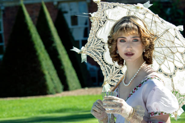 Beautiful lady in regency clothing sitting on lawn in front of stately home holding parasol and drinking sparkling wine Beautiful woman in vintage clothing sitting on lawn in front of stately home holding parasol and drinking sparkling wine regency style stock pictures, royalty-free photos & images