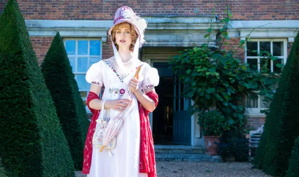 Photo of Beautiful woman in vintage Georgian dress and bonnet standing in garden of stately home