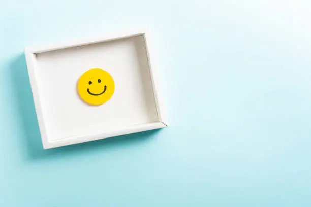Photo of Concept of well-being, well done, feedback, employee recognition award. Happy yellow smiling emoticon face frame hanging on blue background with right empty space for text.
