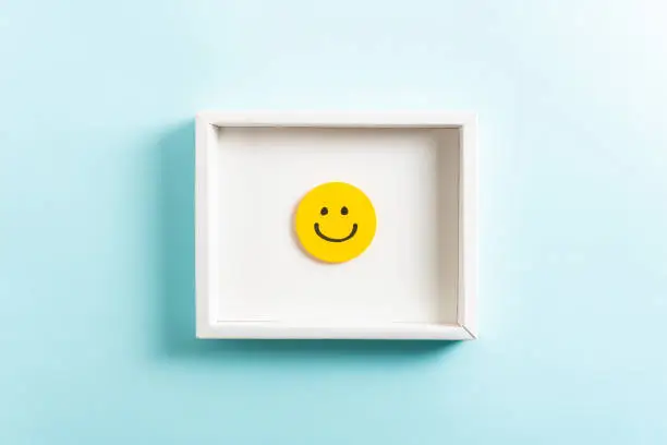 Photo of Happy diploma concept. Concept of well-being, well done, feedback, employee recognition award. Happy yellow smiling emoticon face frame hanging on blue background.