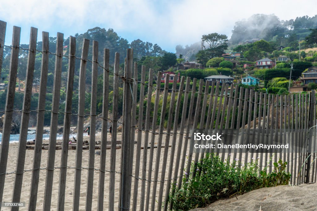 Fence Along the Beach A wooden fence runs along Muir Beach. Low fog over the hillside homes with blue sky peaking out. Beach Stock Photo