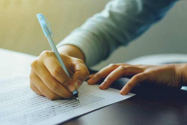 istock Close up of student's hand holds a pen write on answer sheet. Student answers multiple choice questions on wooden table in examination room. 1171332203