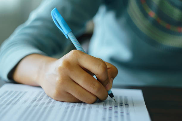 istock Close up of student's hand holds a pen write on answer sheet. Student answers multiple choice questions on wooden table in examination room. 1171333056