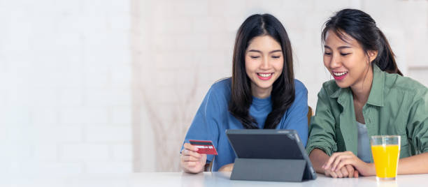 candid of young attractive asian two girls sitting at home holding discount credit card in hand paying for shopping online at tablet smartphone in omni channel concept. asian youth casual lifestyle. - couple laptop computer digital tablet imagens e fotografias de stock