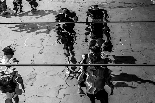 The distorted reflection and shadows of pedestrians walking on a public sidewalk.  The reflection is cast by a metallic canopy that stretches across the sidewalk.  London, England. Monochrome.