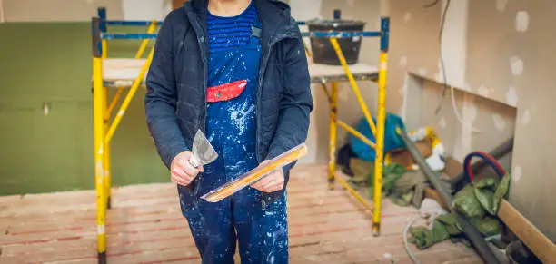 Worker holds a palette-knife for flattening the ceiling from wooden platform in room of apartment is inder construction, remodeling, renovation, overhaul, extension, restoration and reconstruction. Concept of home improvement