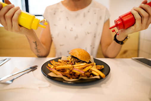 Cropped shot of a woman adding mustard and tomato ketchup to the plate with burger and chips at a restaurant