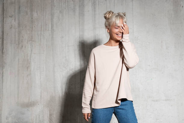 Joyful blonde woman touching head posing in studio Portrait of cute girl wearing powder coloured sweatshirt and blue jeans. Female shyly blushing and covering face with hands on concrete background. Copy space on left side rosy cheeks stock pictures, royalty-free photos & images