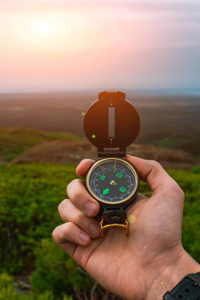 travel, camping, orienteering and navigation concept - black magnetic compass close-up in a man s hand, blurred landscape background - azimuth imagens e fotografias de stock