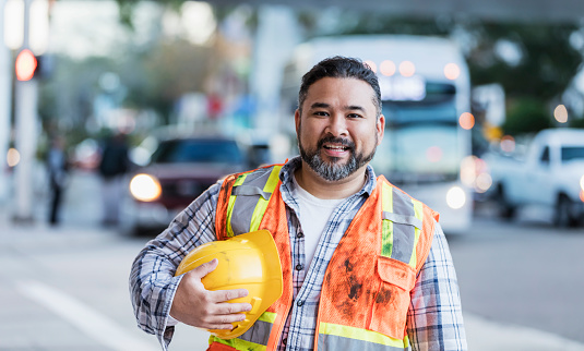 A mid adult Hispanic man in his 30s standing on a city street wearing a reflective vest, holding a hard hat. He is a utility worker or construction worker, looking at the camera.