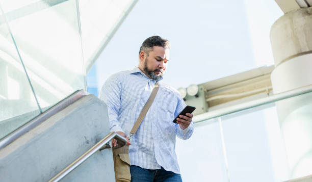 Hispanic man walking, looking at mobile phone A mid adult Hispanic man in his 30s wearing a button down shirt, walking down stairs outside a building, looking down at his mobile phone. shoulder bag stock pictures, royalty-free photos & images