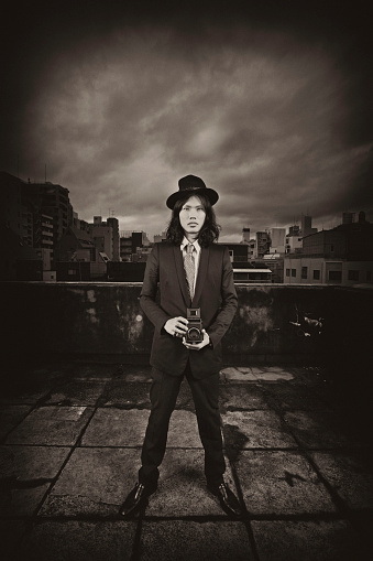 Side profile of a cool young man standing on a roof top with the Tokyo Cityscape in the background.  Male is holding a vintage camera looking straight on towards the camera.  Image is processed in a sepia tone and grain has added.