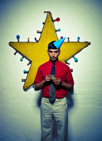 Portrait of a solitary young male standing in front of a wall decoration, wearing a blue party hat and holding a noise maker.