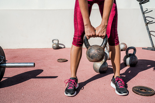 Woman weightlifting kettlebell weight at outdoor fitness gym. Unrecognizable female athlete strength training legs, glutes and back lifting free weights. Closeup of lower body.