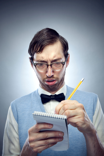 Nerdy IT young man wearing a bow tie, blue sweater vest, and horn rimmed glasses makes a confused, goofy face while writing on a pad of paper with a yellow pencil. Vertical on gray / blue background with copy space.