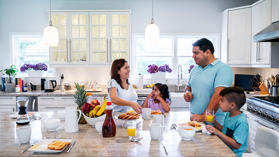 Mature Hispanic parents enjoying breakfast with their young son and daughter at a granite island in their spacious kitchen.