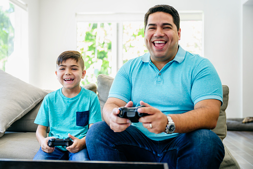Laughing Latin American father and young son sitting on a sofa in the family home and playing video games.