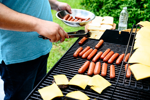 Close-up of mature man barbecuing hot dogs and cheeseburgers on backyard grill for summertime cookout.